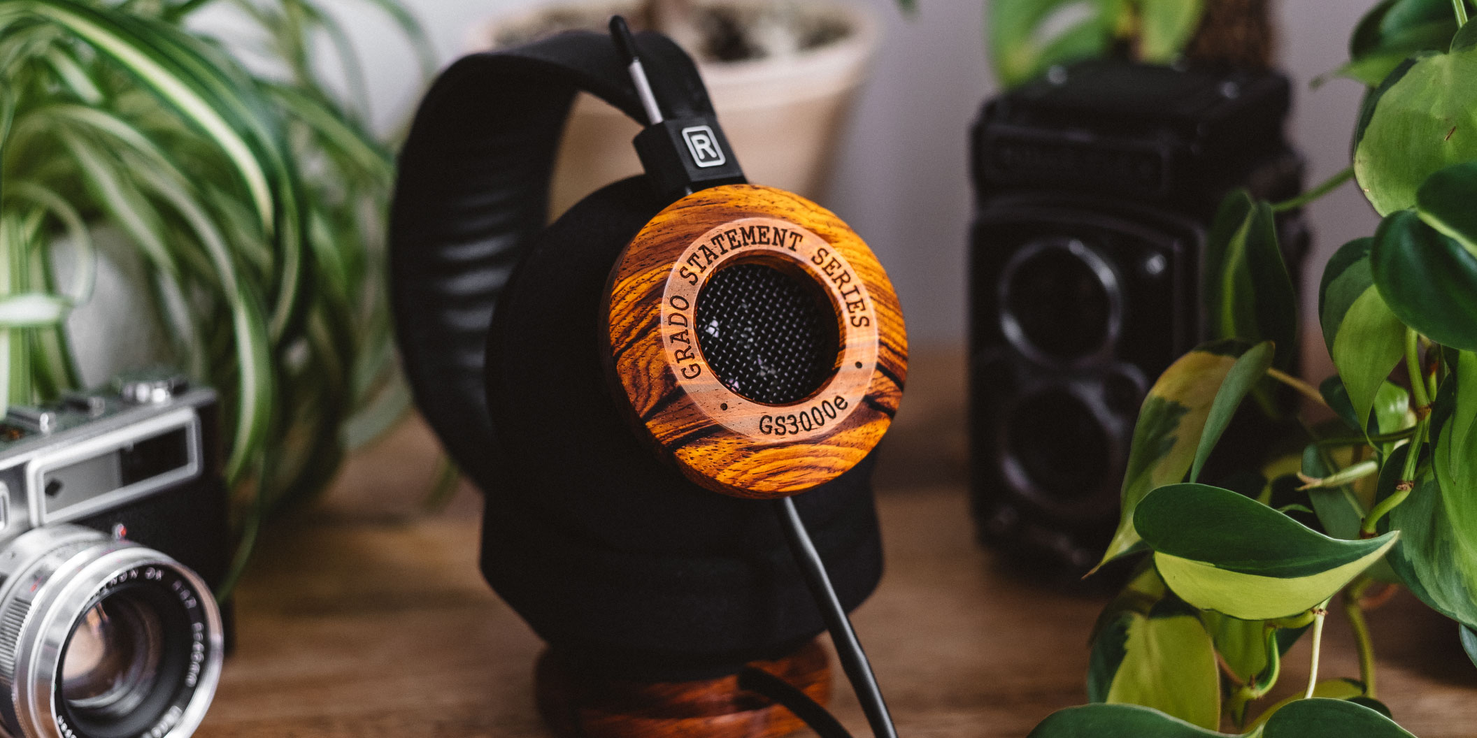 Photo of GS3000e Headphones resting on a stand on a wooden table, surrounded by plants and next to a camera.