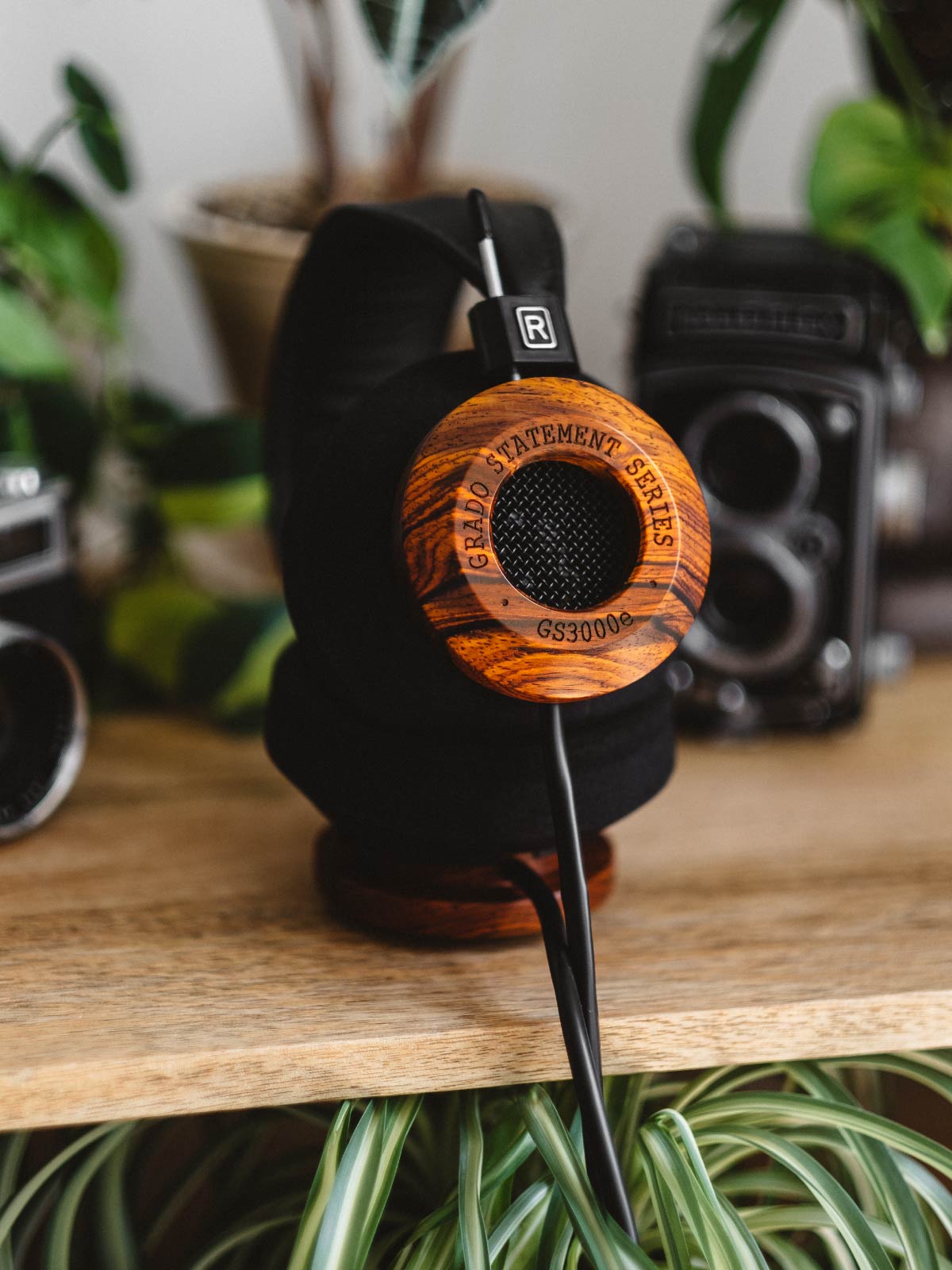 Photo taken from the side of GS3000e headphones resting on a stand on a wooden table surrounded by plants and cameras